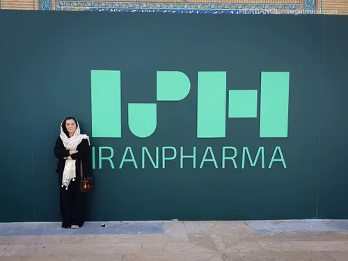 Woman in headscarf standing beside large green IranPharma logo wall
