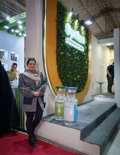 Farvah Fereidounfar in a gray coat stands next to oversized models of pharmaceutical vials labeled ‘Dyston’, in front of a booth with lush green vertical garden backdrop at an exhibition.