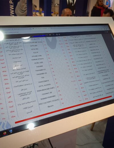 Close-up view of an electronic display screen showing a list of chemical ingredients in both English and Persian, with a reflection of the exhibition hall lights.