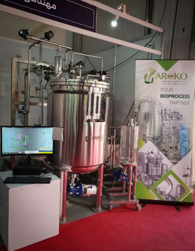 Industrial bio-processing stainless steel equipment with various pipes and valves, displayed next to a poster reading ‘AROKO - YOUR BIOPROCESS PARTNER’ at a trade show.