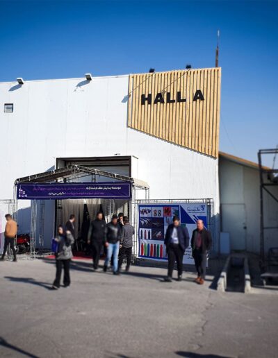 Entrance to Iran Cosmetica exhibition hall named ‘HALL A’ with people entering and exiting, flanked by promotional banners, under a clear blue sky.