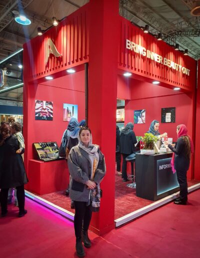 Farvah Fereidounfar posing in front of a red exhibition stand with the slogan ‘BRING INNER BEAUTY OUT’, showcasing beauty products, with attendees and staff in the background.