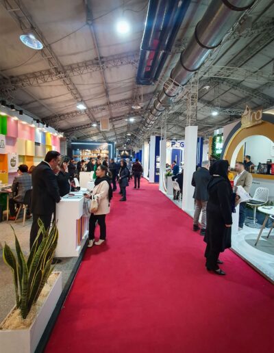 Busy Iran Cosmetica exhibition hall with a red carpet, various booths with brand logos, and visitors engaging with the exhibits, under bright ceiling lights.