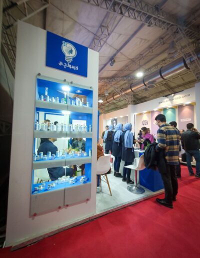 Iran Cosmetica indoor exhibition space with a long red carpet leading past various colorful cosmetic and pharmaceutical booths, busy with visitors and staff. “Formoulchi” and “فرمولچی”.