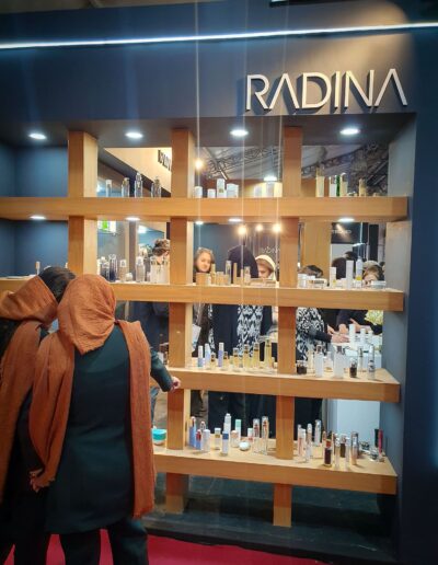 A well-lit exhibition booth with the ‘RADINA’ brand prominently displayed above a wooden shelf showcasing a variety of beauty and skincare products, with visitors engaging with the products.
