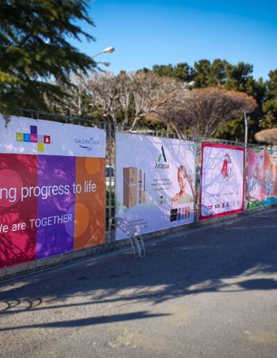 A row of vibrant, colorful advertising banners displayed on a fence along a tree-lined street, promoting various brands with messages of progress and unity.