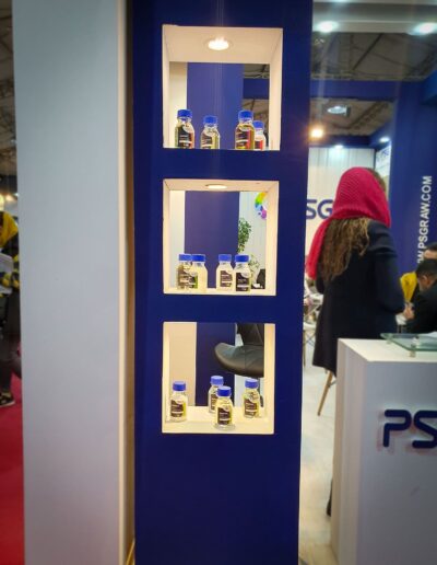 A booth at an exhibition displaying blue and white cosmetic products on white shelves, with visitors and staff in attendance, under overhead lighting.