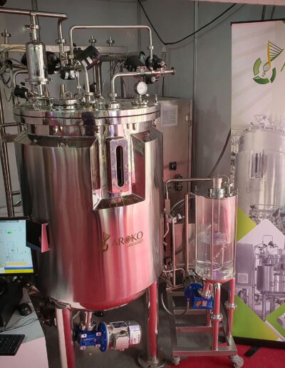 Industrial bio-processing stainless steel equipment with various pipes and valves, displayed next to a poster reading 'AROKO - YOUR BIOPROCESS PARTNER' at a trade show.
