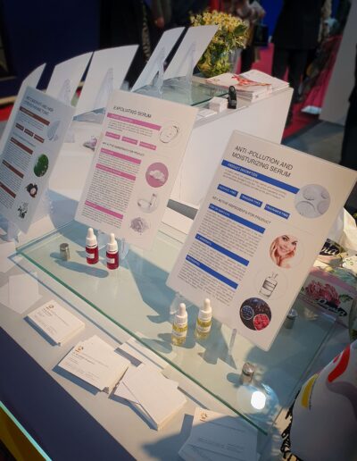 An exhibition display with detailed informational cards about exfoliating and anti-pollution serums, alongside small sample bottles arranged on a glass table.
