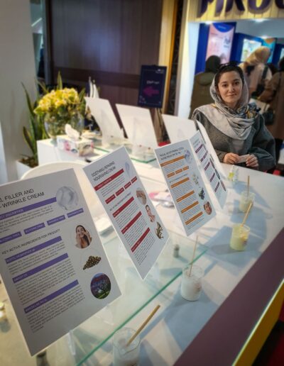 Farvah Fereidounfar in a gray coat and scarf stands behind a display of cosmetic products with information panels explaining the benefits of each skincare item.