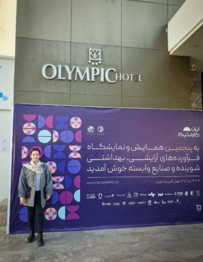 Farvah Fereidounfar in a grey coat and scarf standing in front of a large purple promotional banner outside the Olympic Hotel, featuring cosmetic product advertisements.