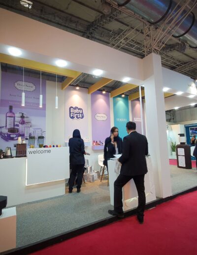 A modern and spacious exhibition booth with white and lilac branding for 'Baby First' and 'PRIME', showcasing beauty and healthcare products with attendees browsing.