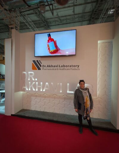 Farvah Fereidounfar in front of the 'Dr. Akhavi Laboratory' exhibition stand, which features white branding on a red background and a promotional video screen above.